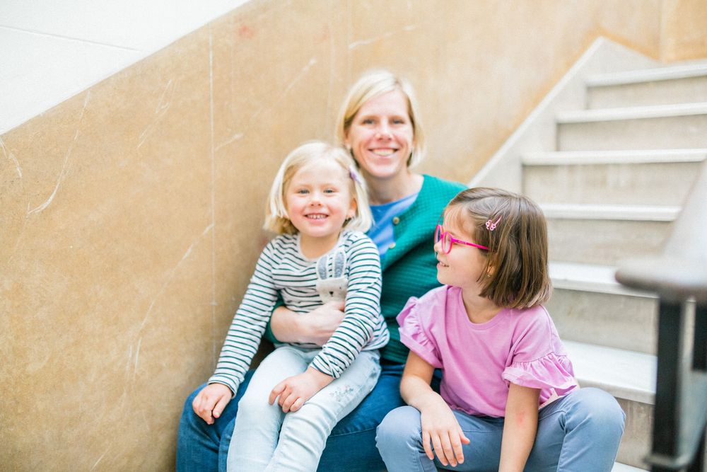 Three people sit together on stairs against a beige wall in a casual family portrait.