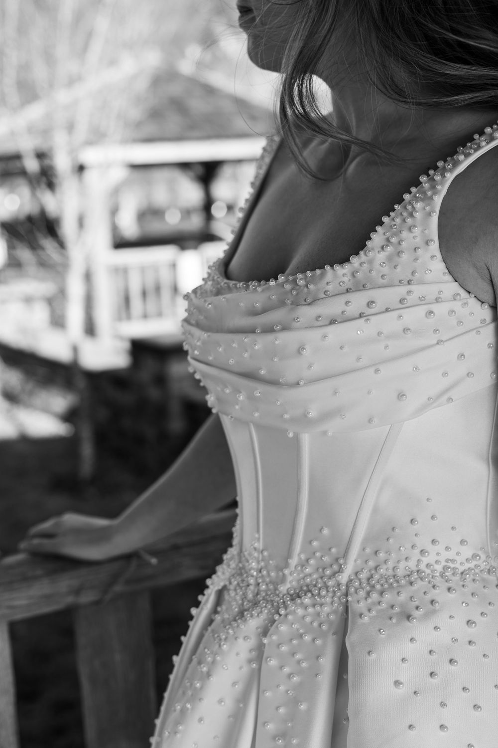 Black and white close-up of a bride in a pearl-embellished wedding gown standing on a wooden balcony.