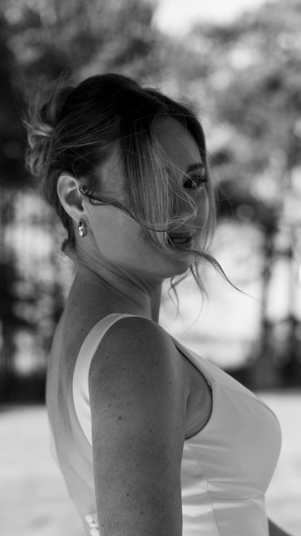 Black and white portrait of a woman in a white dress with wind-blown hair posing outdoors among trees.