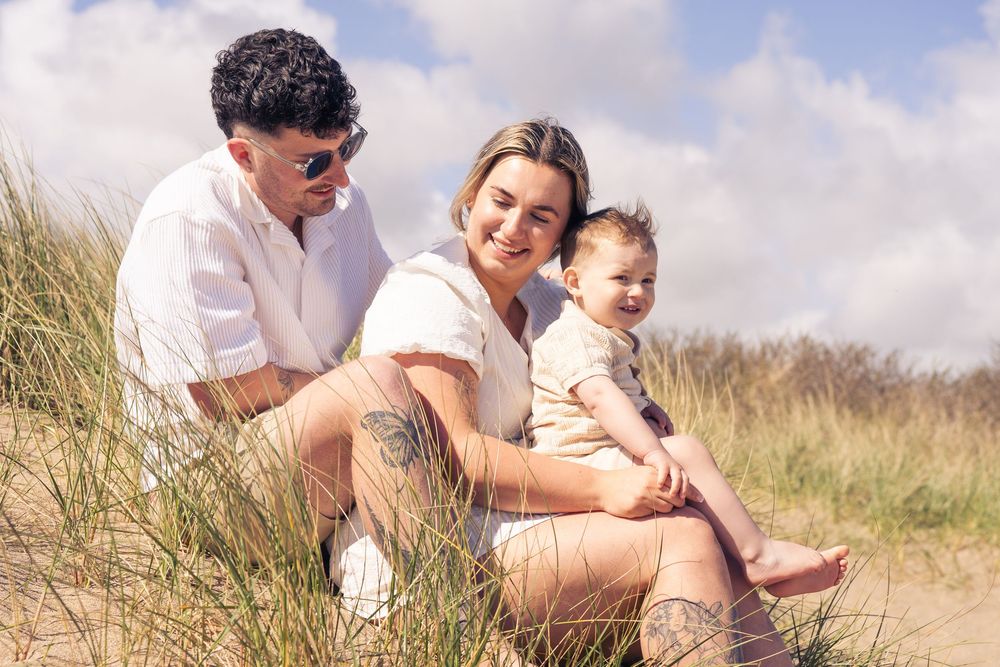 Family of three sitting on sandy dunes, parents smiling with baby on a sunny day near coastal grass.