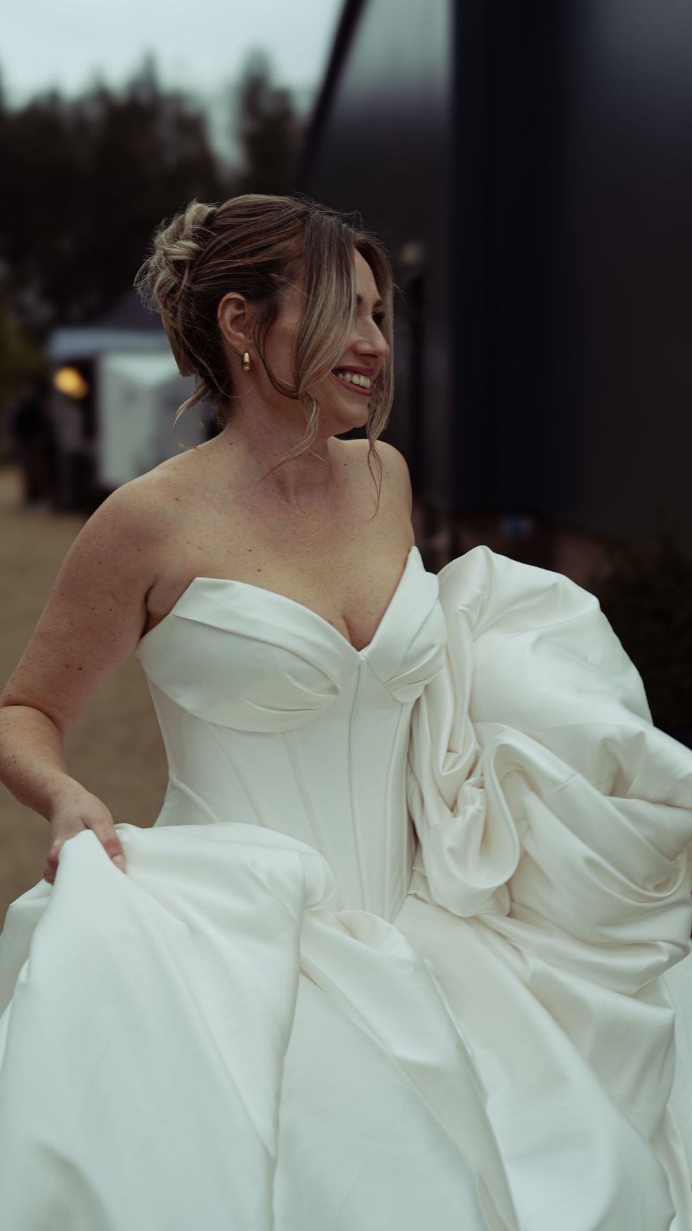 Smiling bride in a strapless white ballgown wedding dress with a large bow detail outdoors.