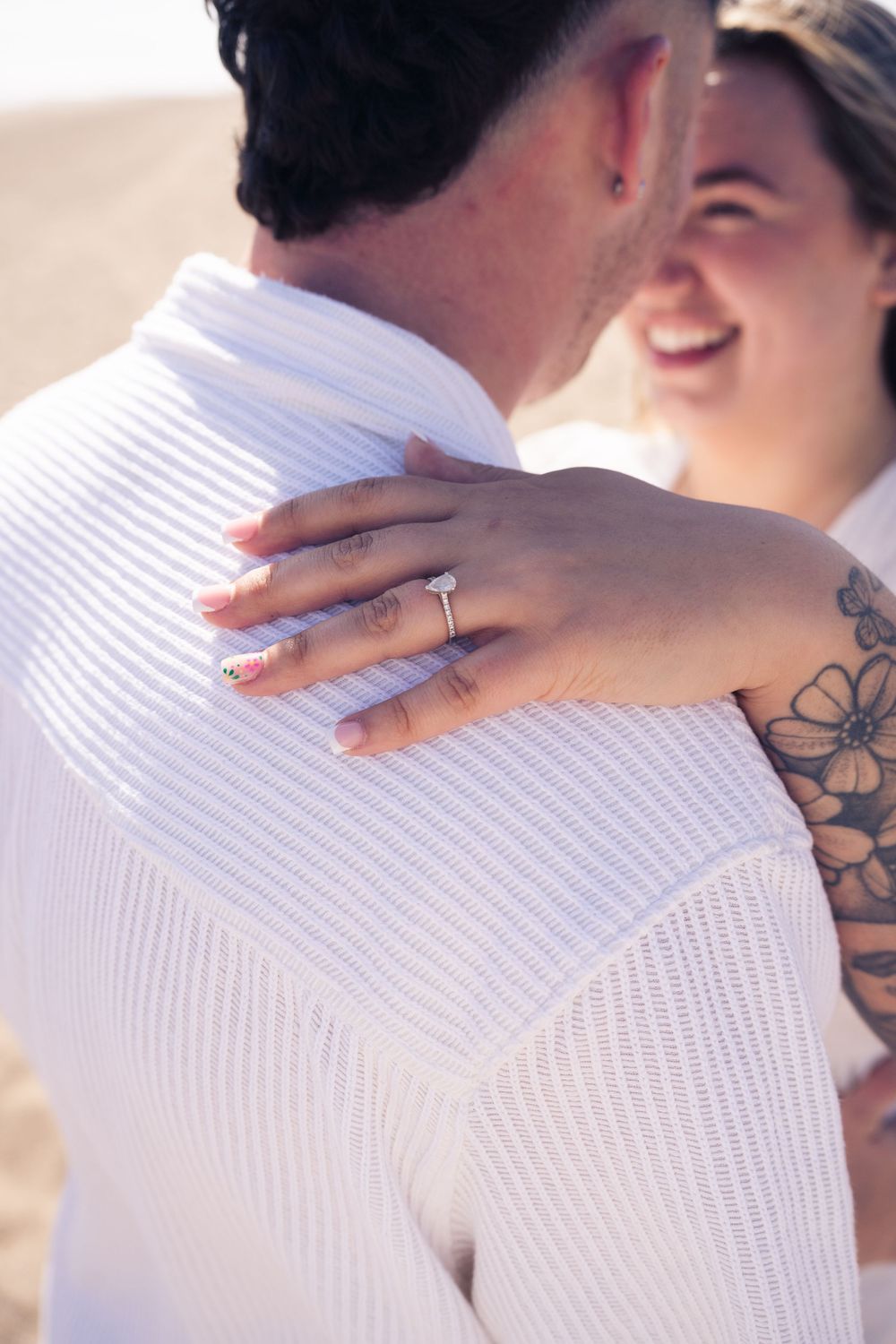 A smiling woman with tattoos hugs a man, showcasing her engagement ring on his shoulder at the beach.
