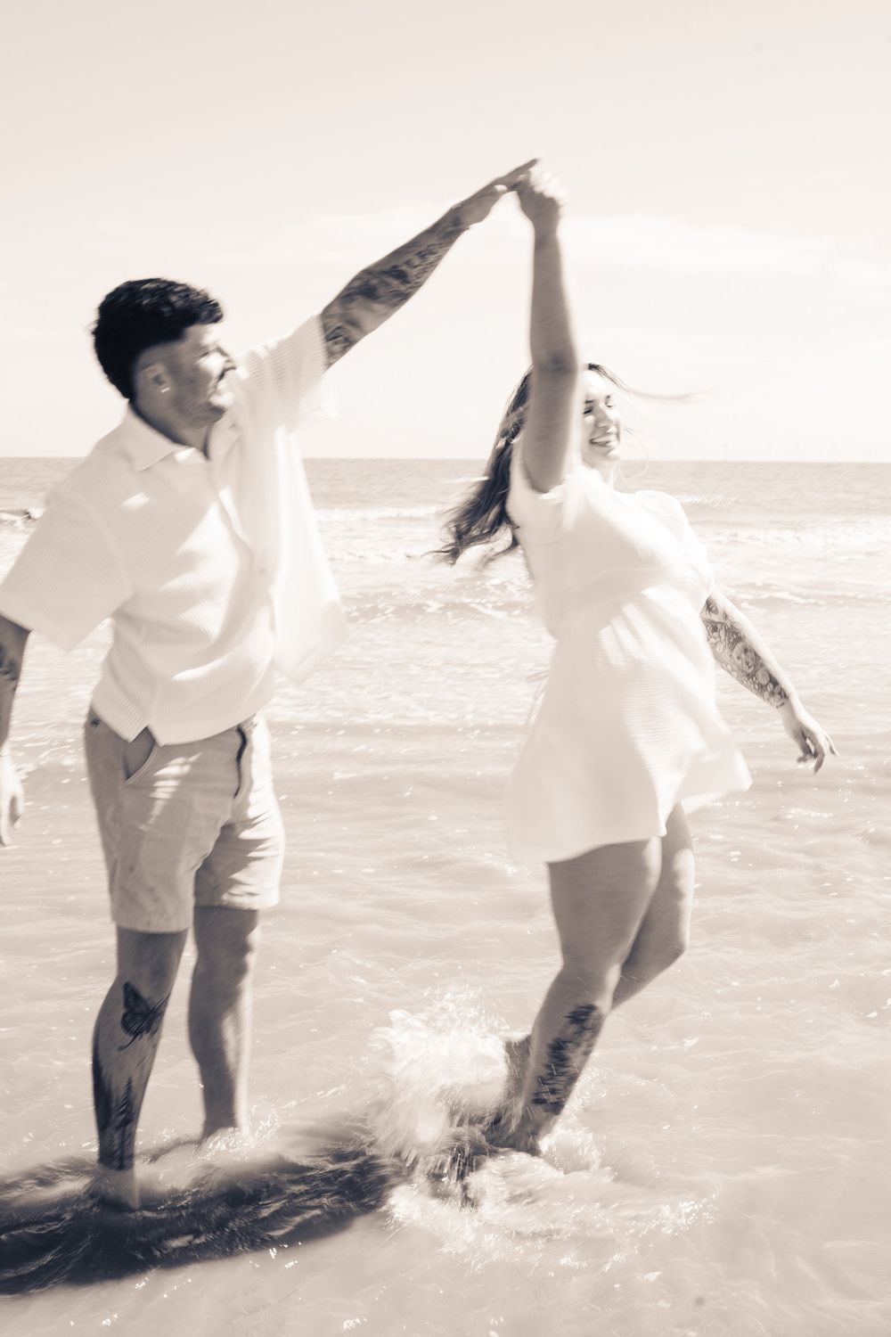 Couple dancing joyfully in ocean waves at beach, wearing white outfits with tattoos, sepia toned photo.