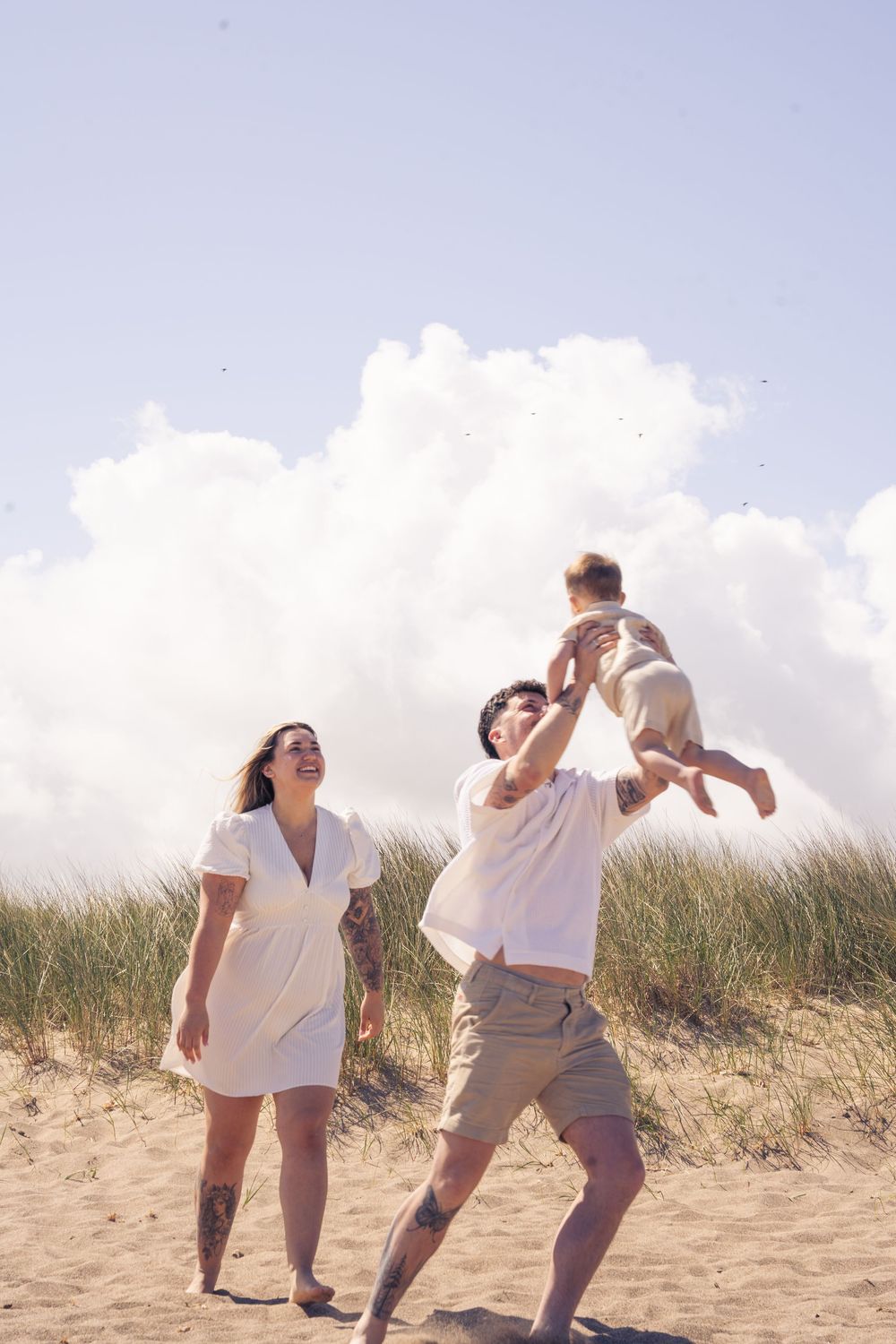 Family playing on sandy beach, father tossing baby in air while mother laughs, with grass dunes and clouds behind.