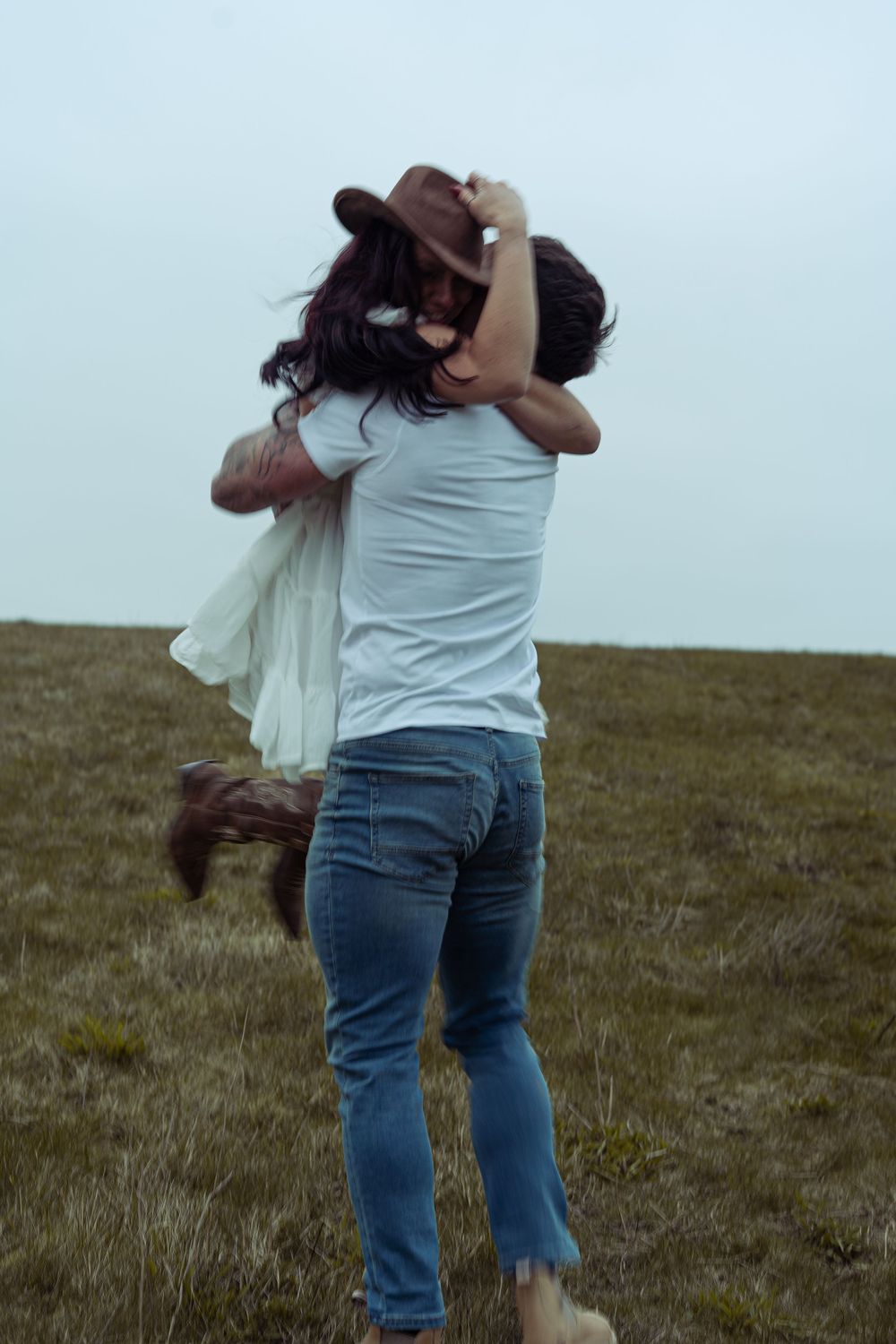 A couple embracing and spinning together in a grassy field, the woman wearing a white dress and cowboy hat.