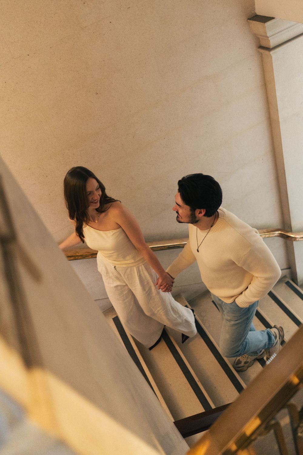 Couple holding hands on elegant staircase, man in cream sweater and jeans, woman in white outfit, sharing a romantic moment.