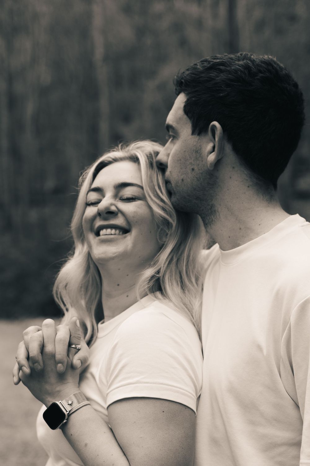 Black and white photo of a laughing woman being kissed on the cheek by her partner outdoors.