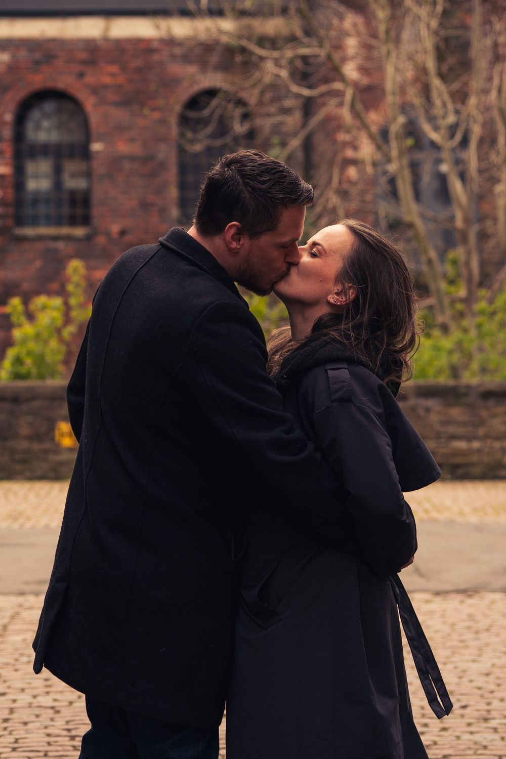 Couple sharing a romantic kiss outdoors in winter coats, with a brick building and bare trees in the background.