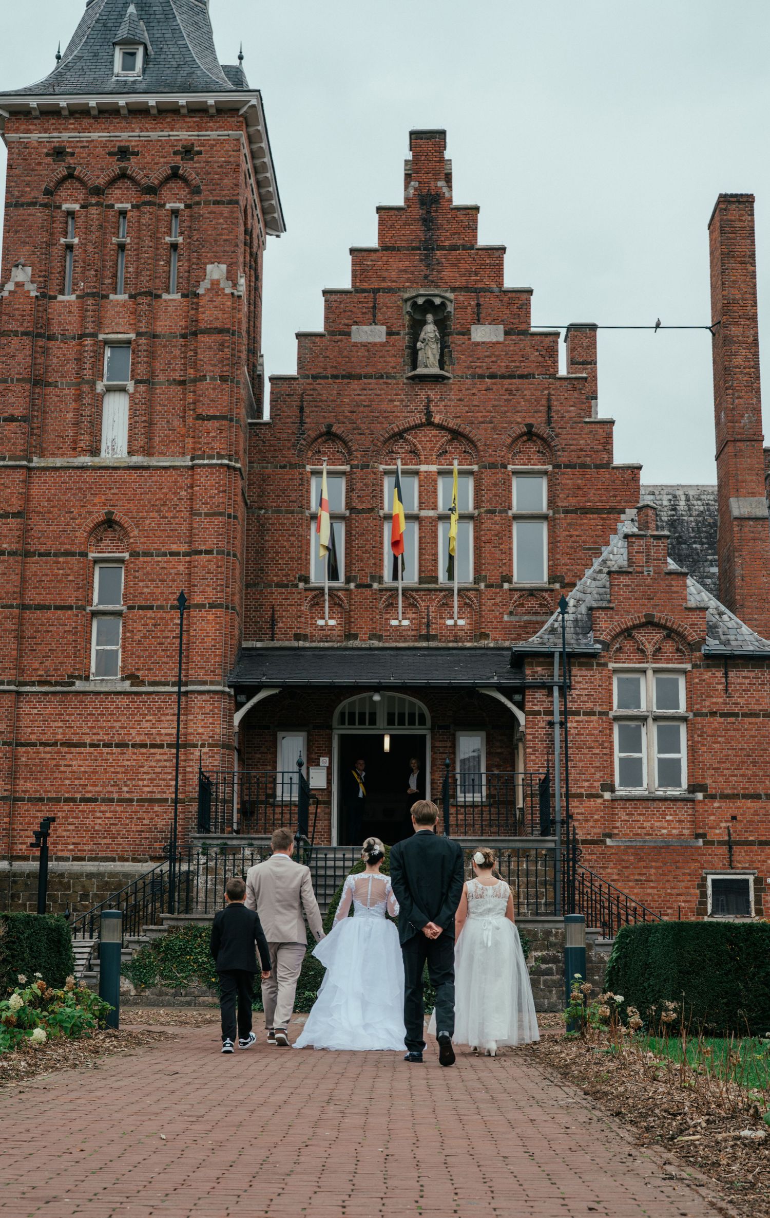 Wedding party walking toward a grand red brick building with stepped gable roof and colorful flags on a cloudy day.