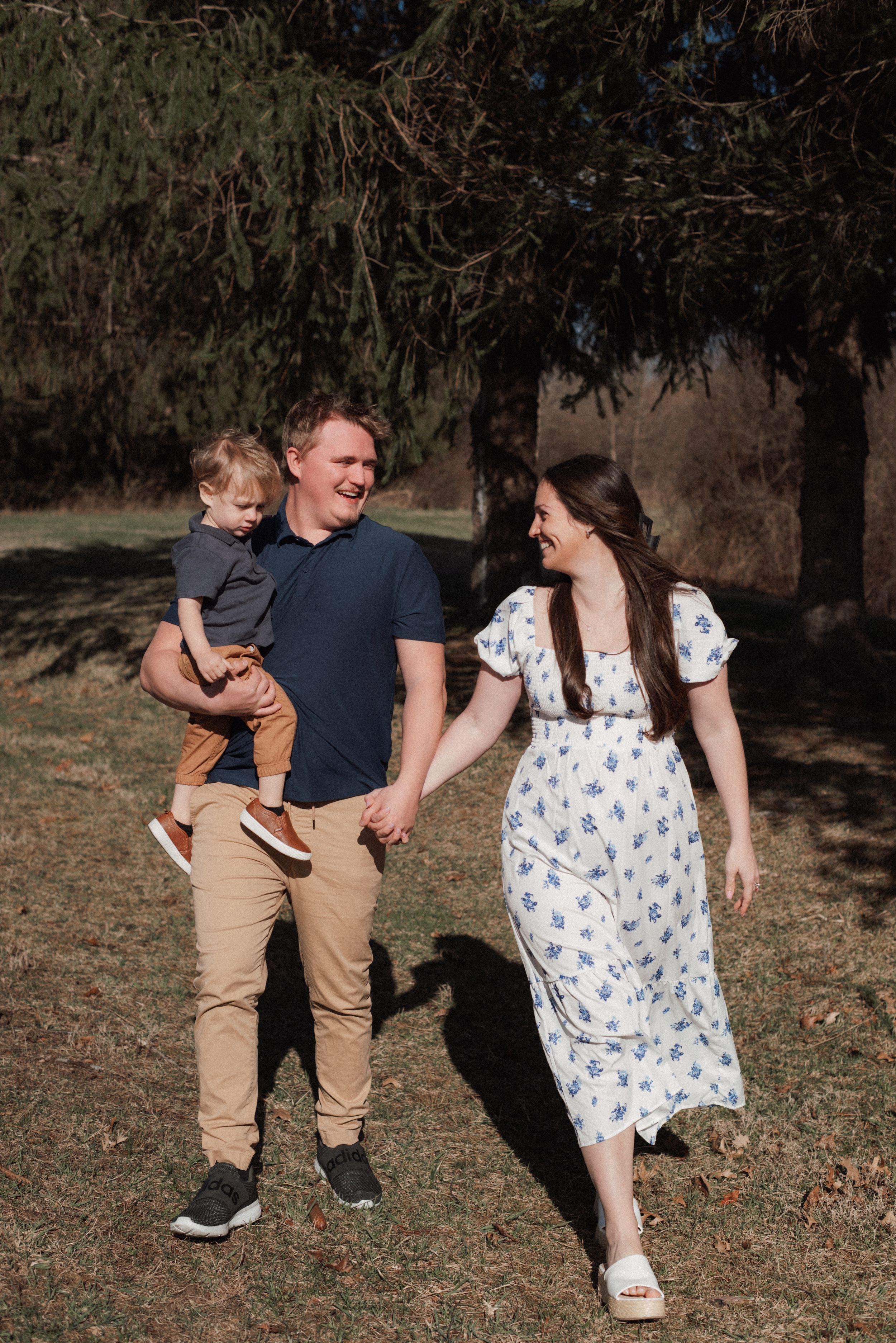 A happy family of three walks outdoors, man holding toddler while pregnant woman in floral dress smiles, near pine trees.