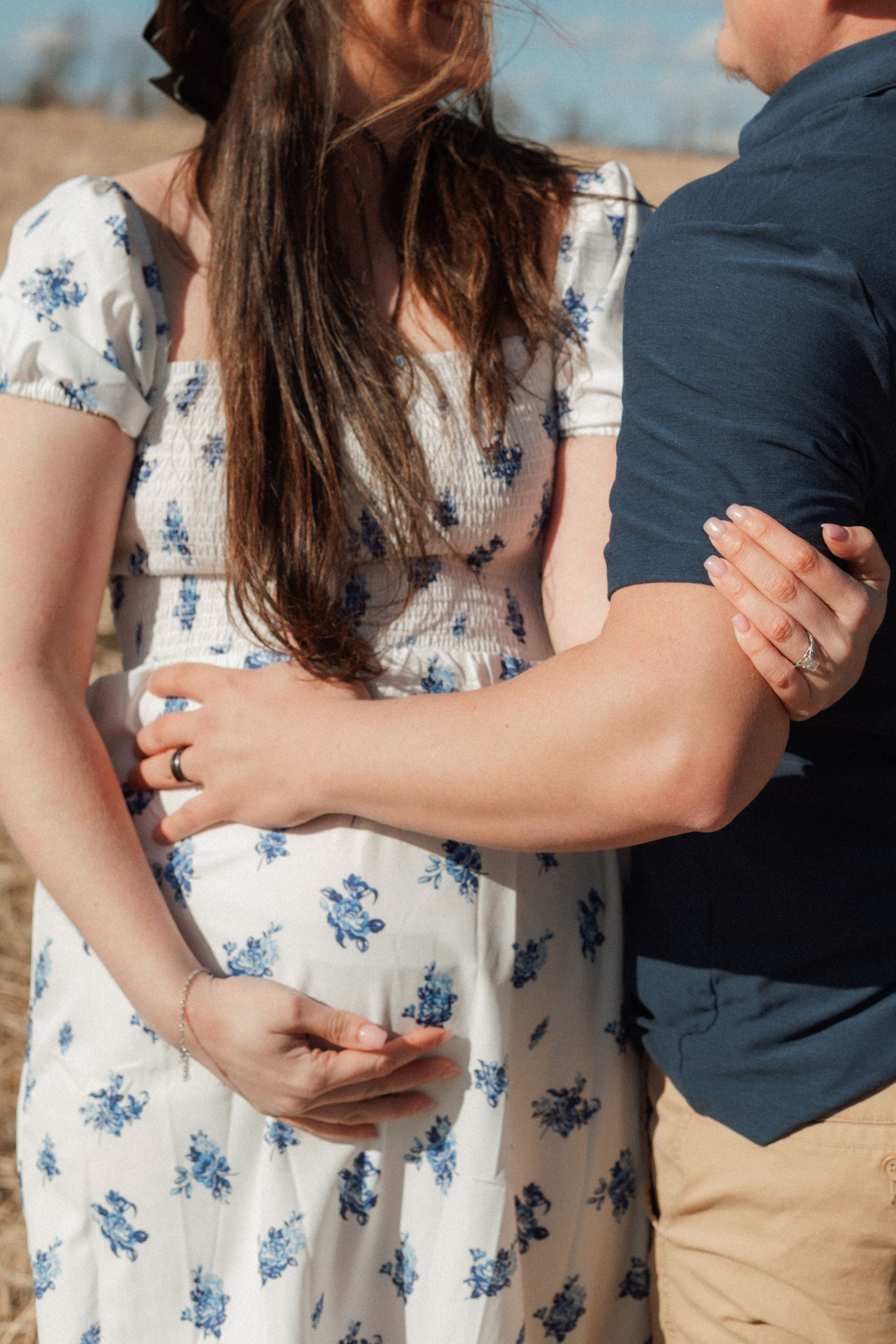 Pregnant woman in floral dress embraced by partner outdoors in sunny field, showing baby bump.