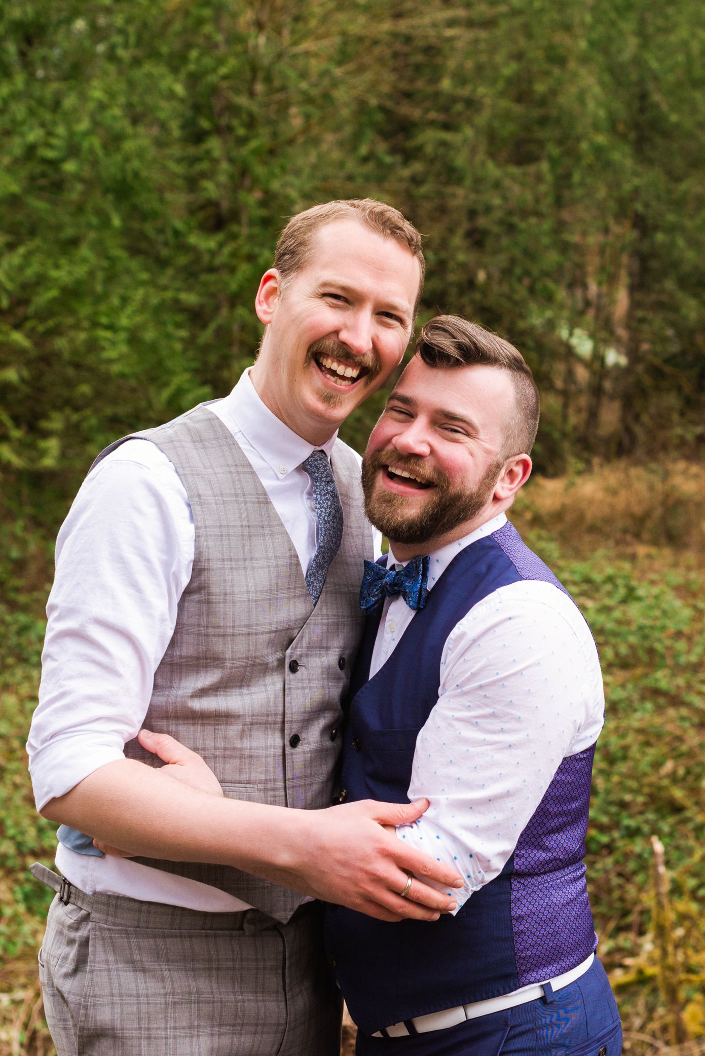 Two grooms in vests embrace and laugh joyfully outdoors during their wedding celebration.