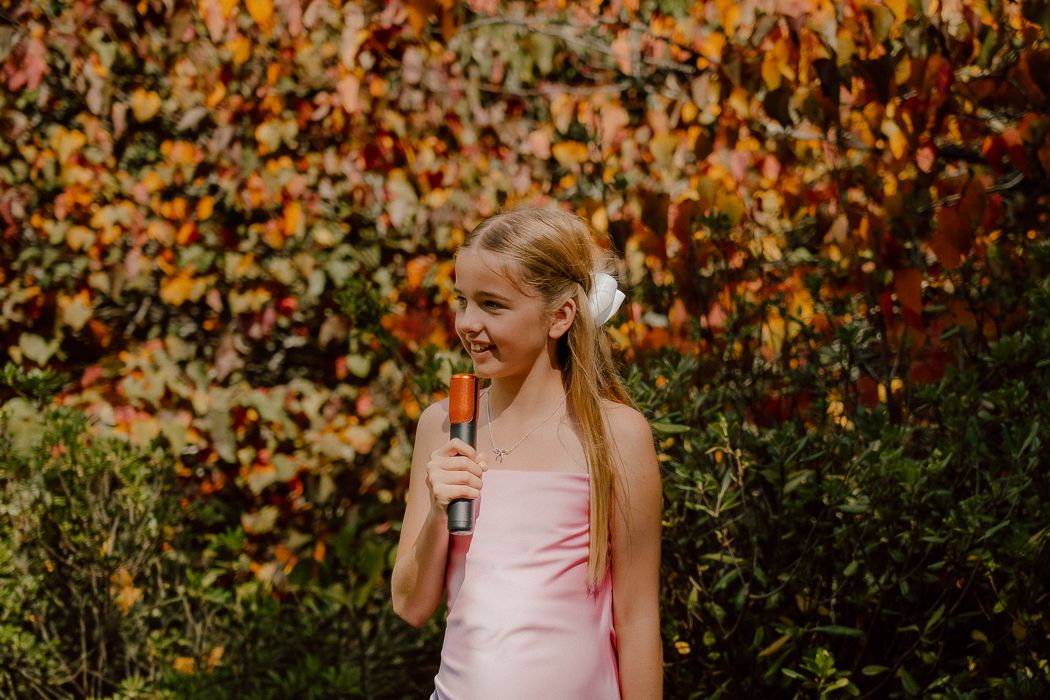 Young girl in pink dress holding microphone, smiling outdoors with colorful autumn foliage background.
