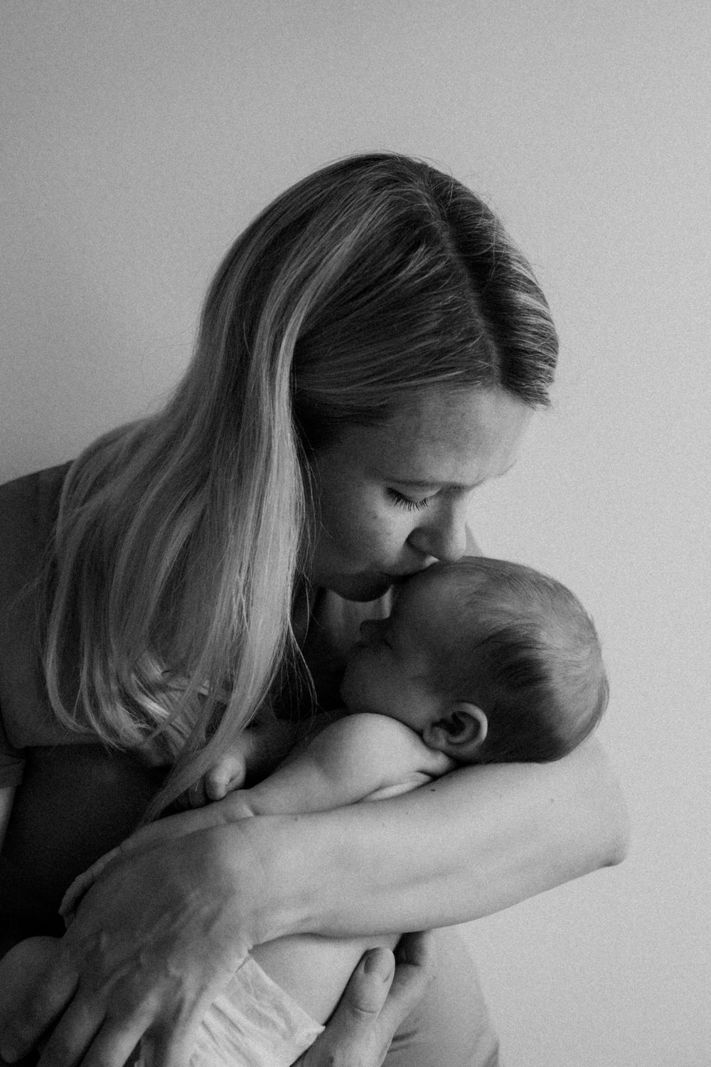 Vastasyntynyt vauva äidin sylissä perheen kotona postpartum- ja vauvakuvauksessa. Äiti pussaa vauvaa otsalle ja katsoo rakastavasti.
