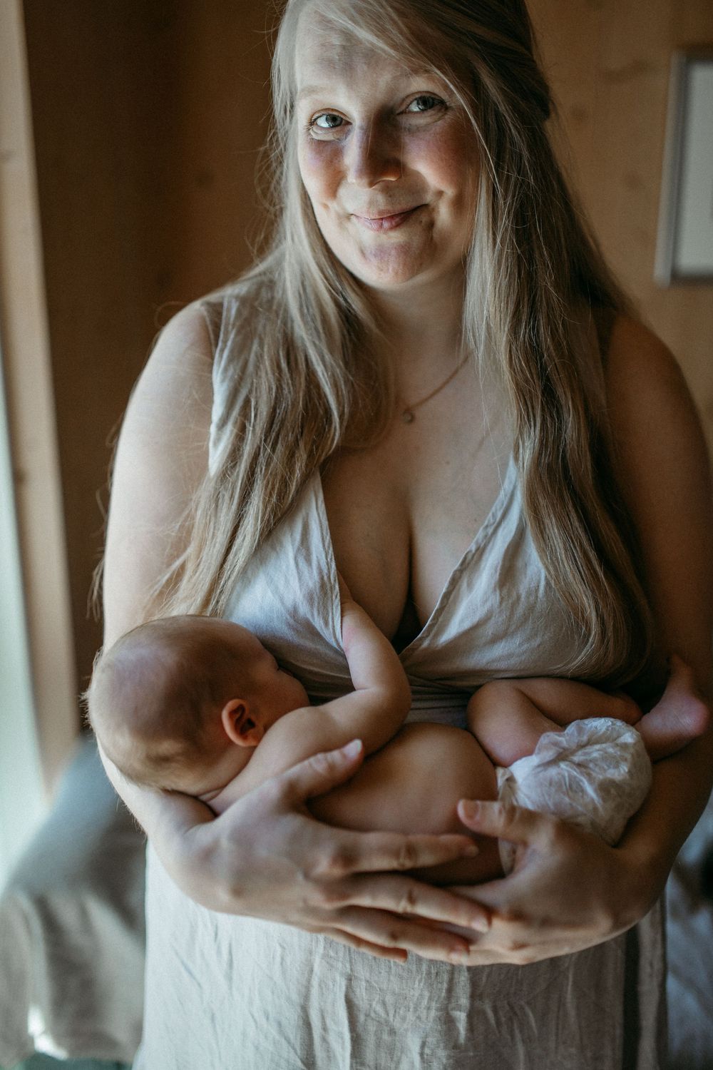 Vastasyntynyt vauva nukkuu äidin rintaa vasten vaippasiltaan perheen kotona postpartum- ja vauvakuvauksessa.Äiti katsoo kameraan onnellisena