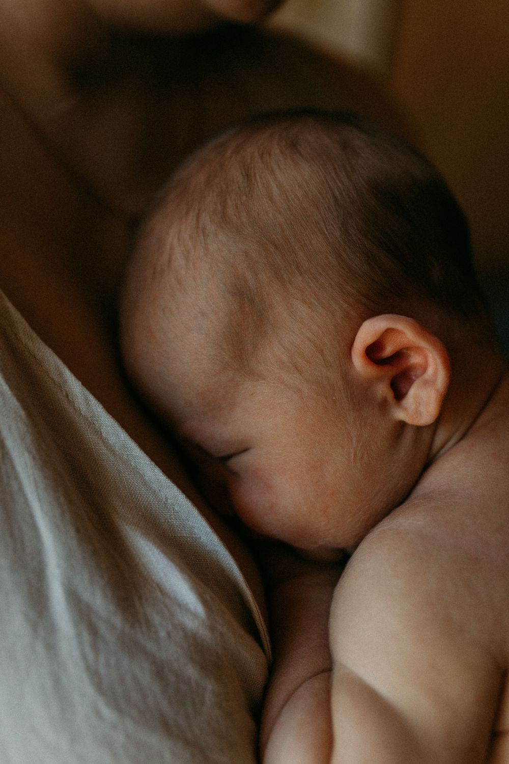 Lähikuva, jossa vastasyntynyt vauva nukkuu äidin rintaa vasten vaippasiltaan perheen kotona postpartum- ja vauvakuvauksessa.