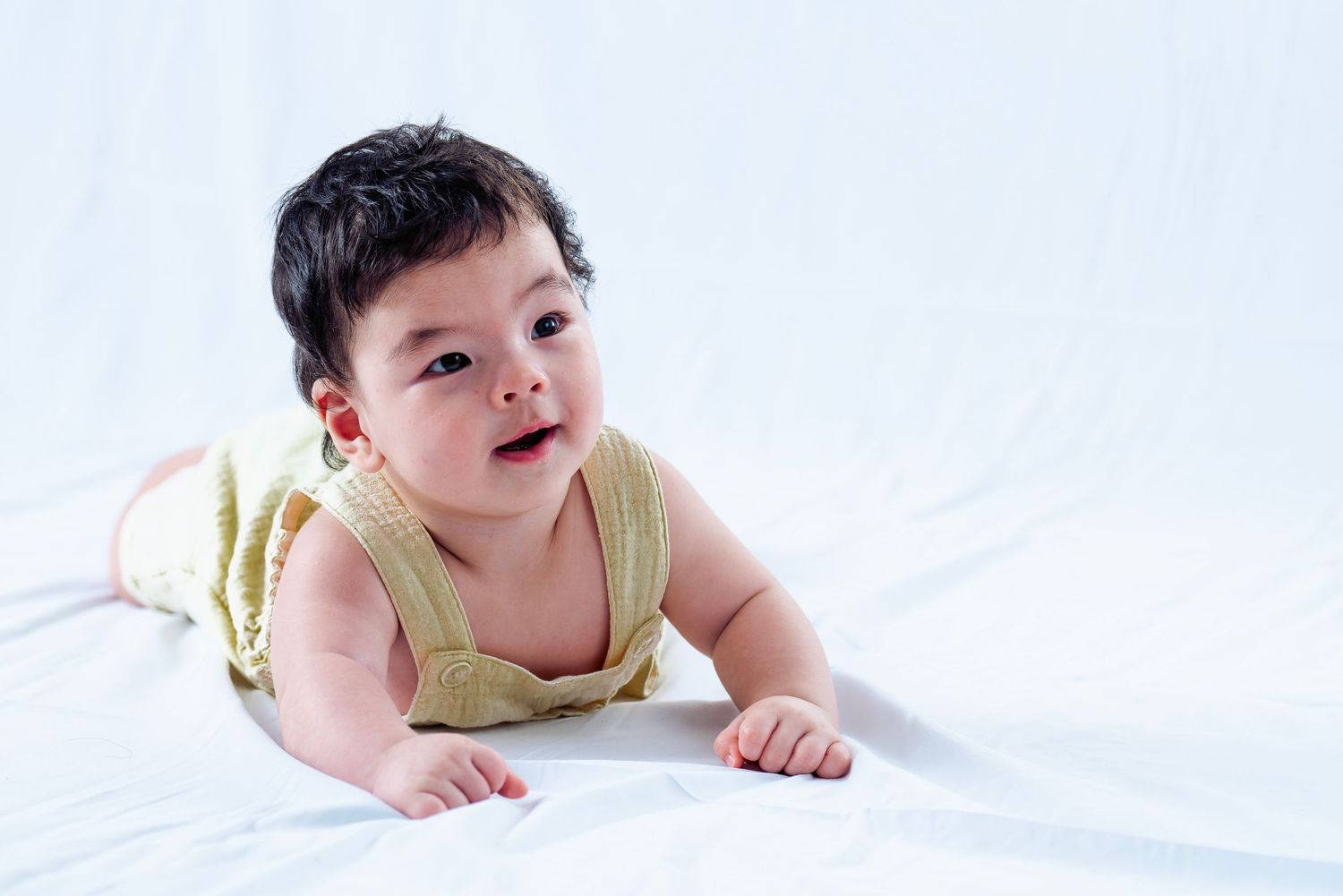 Happy baby in yellow outfit crawling on white sheets.