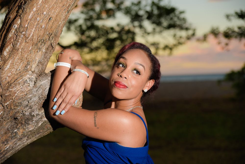 A person in a blue dress poses against a tree trunk during golden hour sunset lighting.
