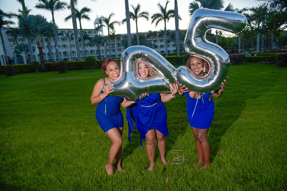Three women in matching blue dresses pose playfully with silver number balloons on a grassy lawn.