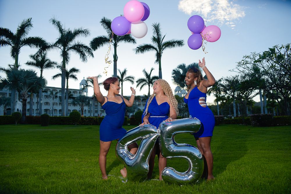 Three people in blue dresses celebrate with silver number balloons and colorful balloons on a palm-lined lawn at dusk.