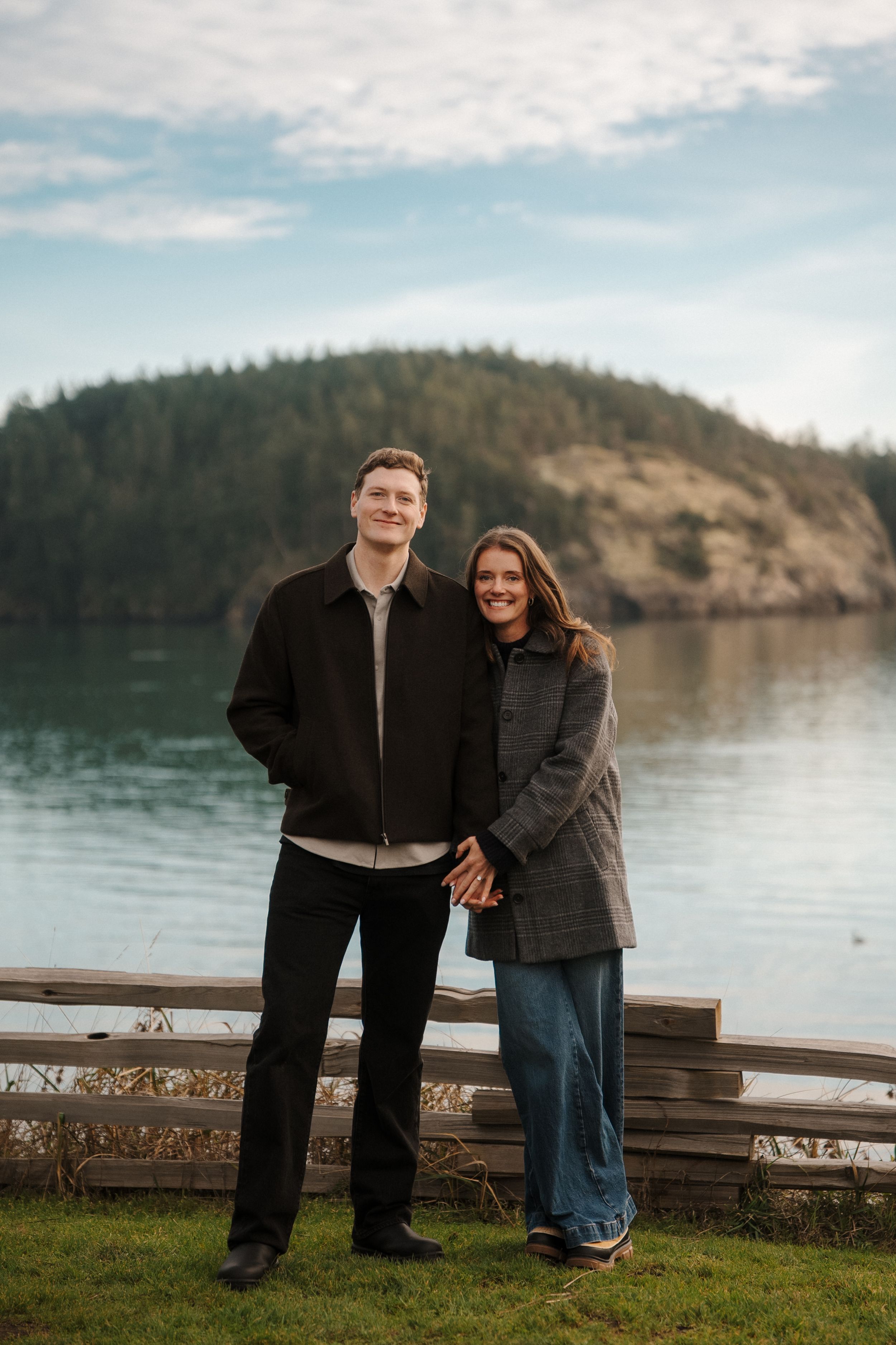 A couple enjoys a romantic moment by the serene waters of a lake with evergreen-covered hills in the background.