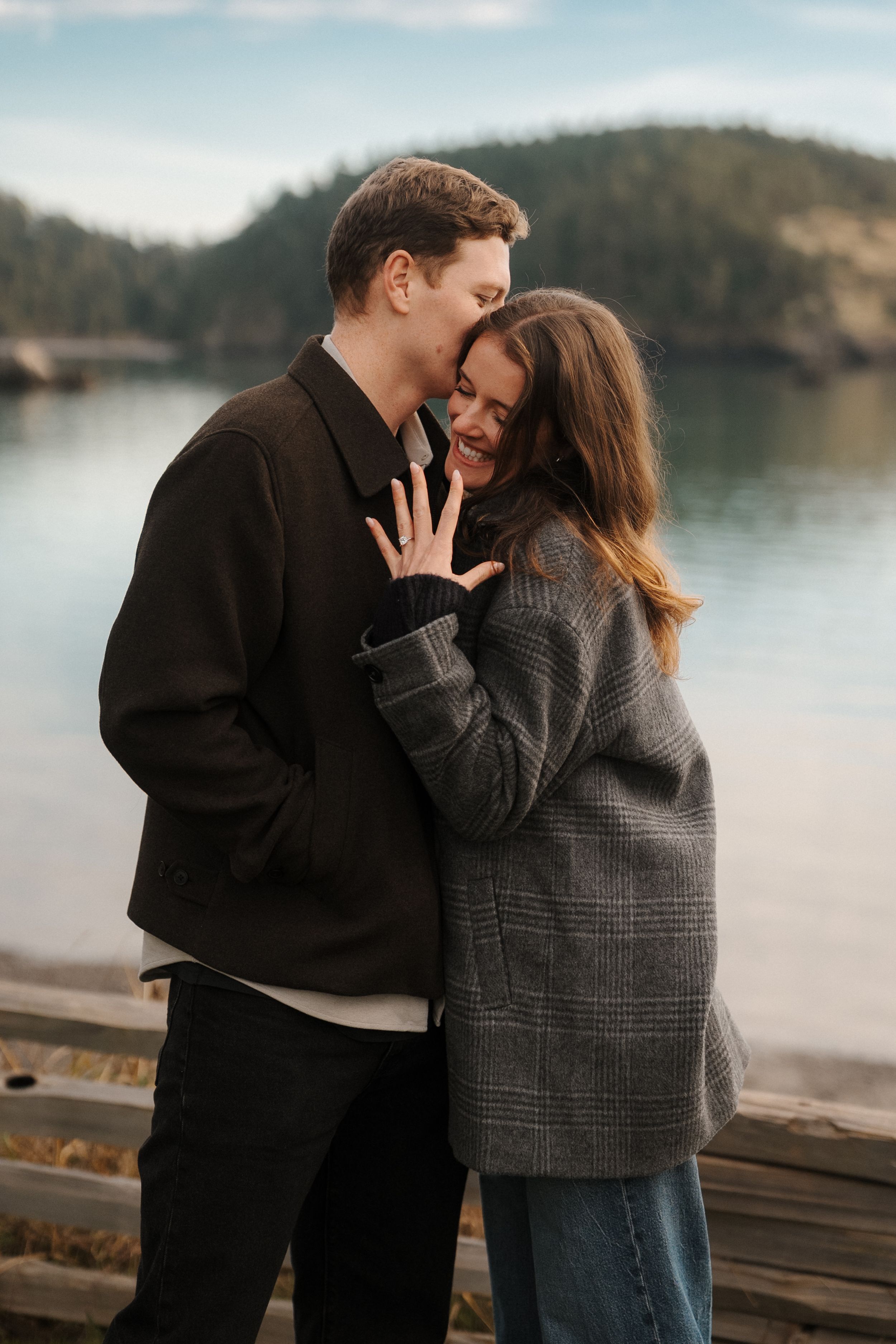 A couple shares an intimate moment while embracing on a dock by a serene lake with mountains in the background.