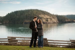 Two people stand together at a scenic overlook by a tranquil lake surrounded by forested hills on a cloudy day.