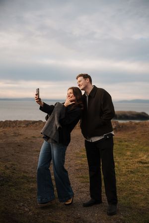 A couple shares a playful moment together while taking selfies on a hilltop against a cloudy sky.