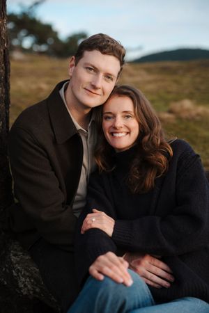 A couple in black sweaters share an intimate moment while sitting outdoors on a rocky terrain.