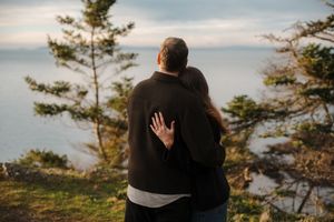A couple embraces while looking out over a scenic coastal viewpoint with evergreen trees at sunset.