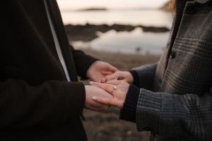 Two people hold hands tenderly at sunset with a blurred waterfront background.