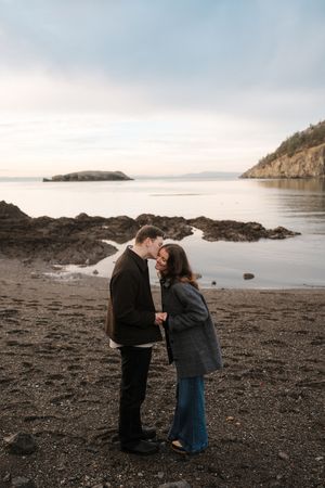 A couple shares an intimate moment on a rocky beach at sunset with mountains and water in the background.
