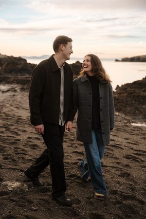 A couple walks hand in hand along a rocky beach at sunset, sharing a joyful moment together.