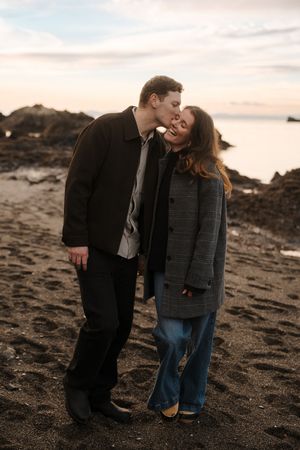 A couple shares an intimate moment on a rocky beach at sunset as they embrace on the dark volcanic sand.