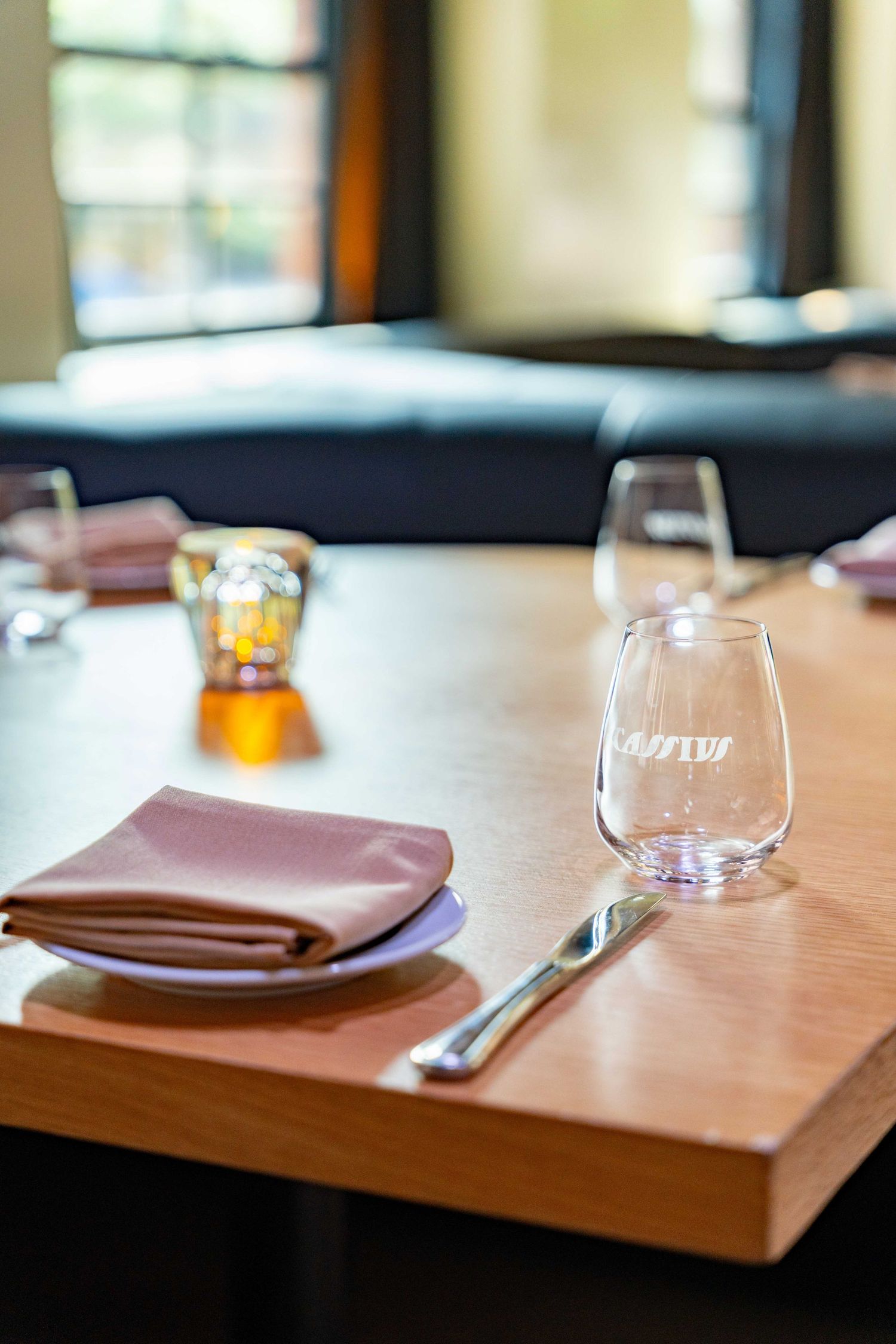 A restaurant table setting with a wine glass, brown napkin, and cutlery on a wooden surface in soft lighting.