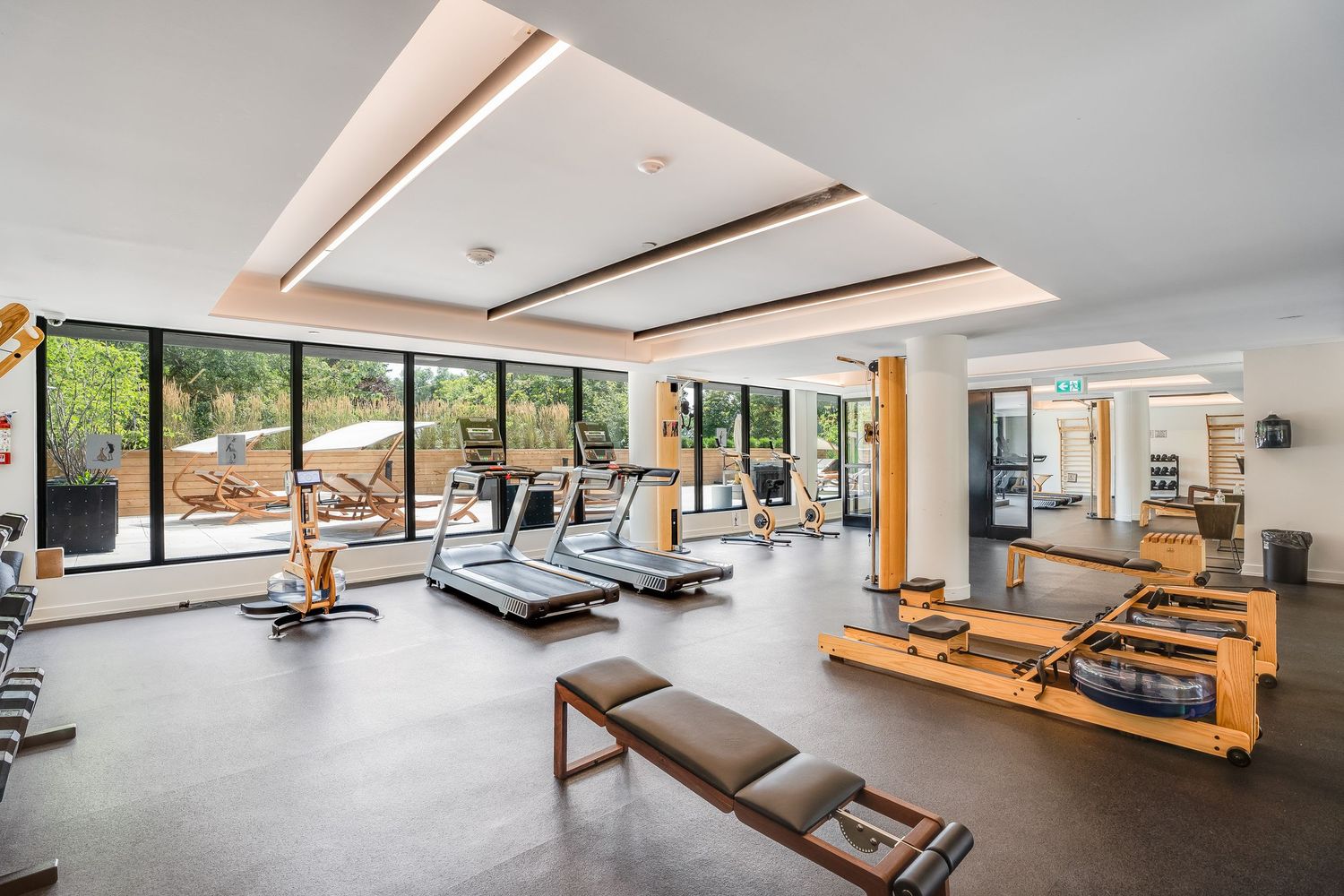 Modern fitness studio with large windows, wooden floors, and exercise equipment arranged along mirrored walls.