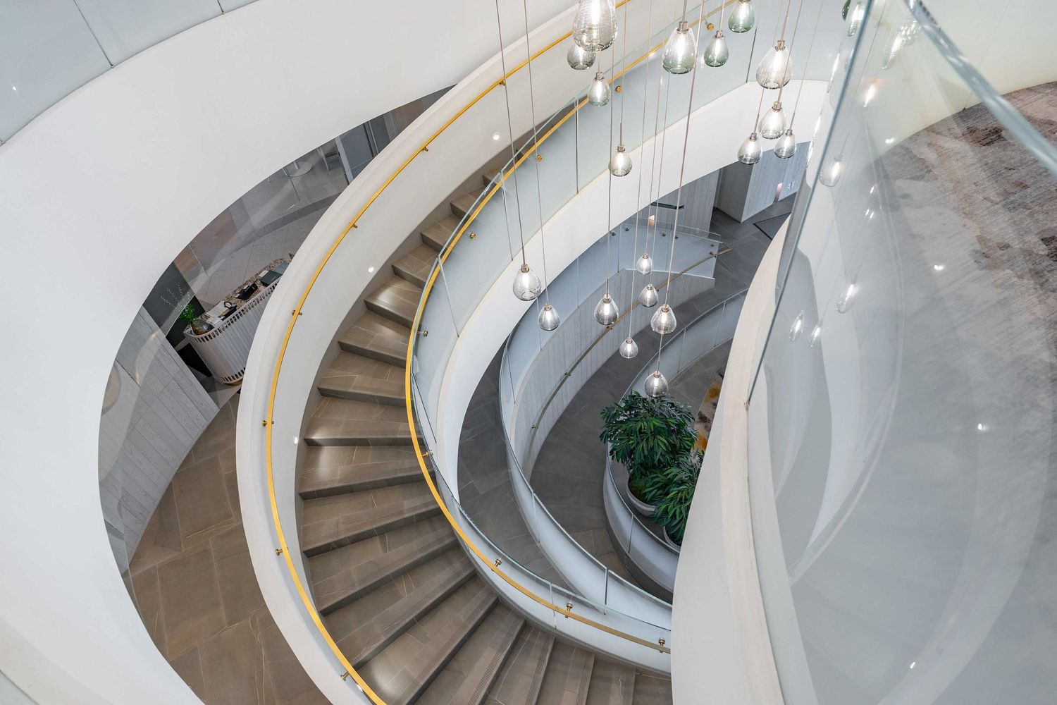Curved white staircase with glass balustrades and pendant lighting viewed from above in a contemporary interior space.