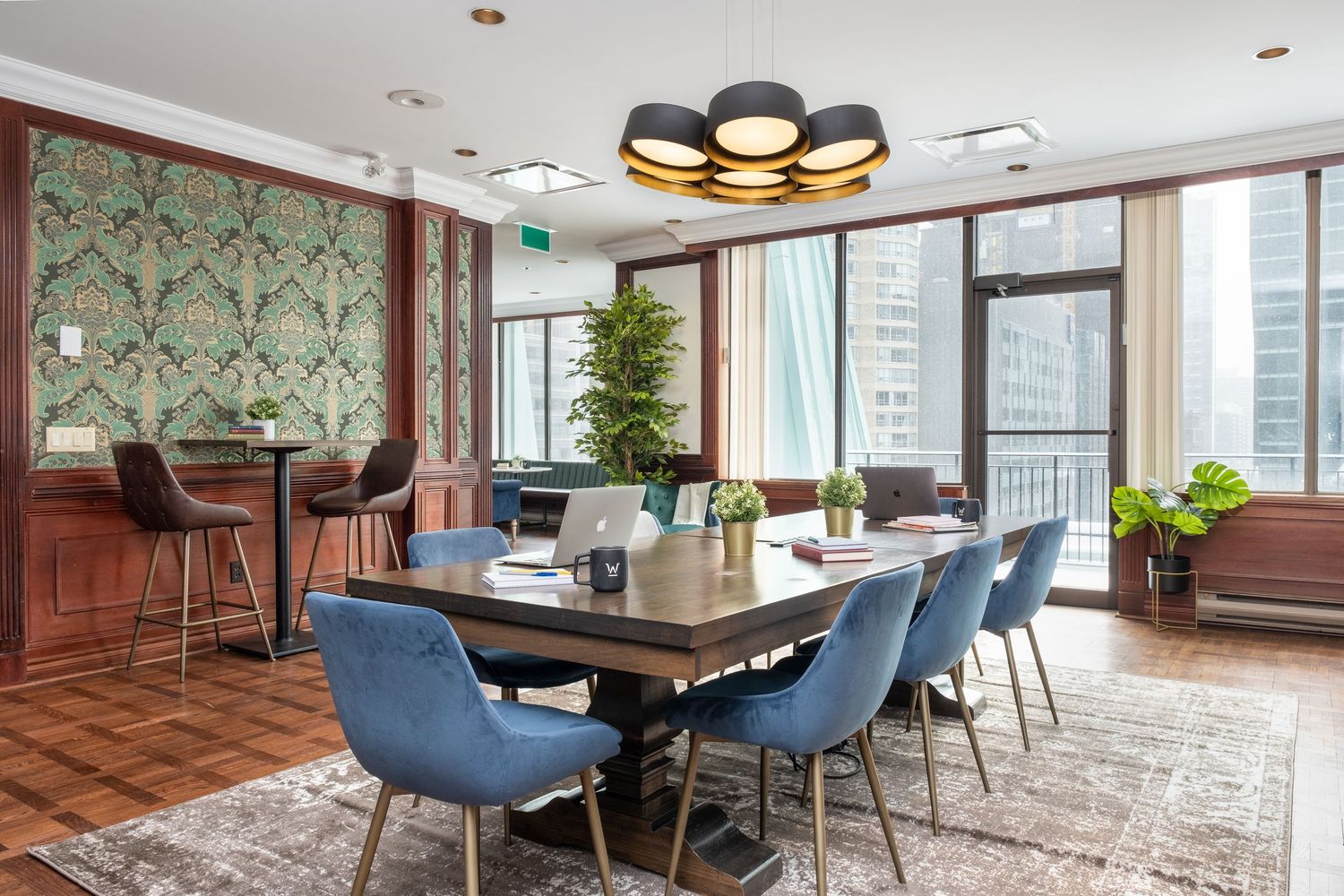 Modern open concept apartment with blue velvet chairs and floor-to-ceiling windows offering city skyline views.