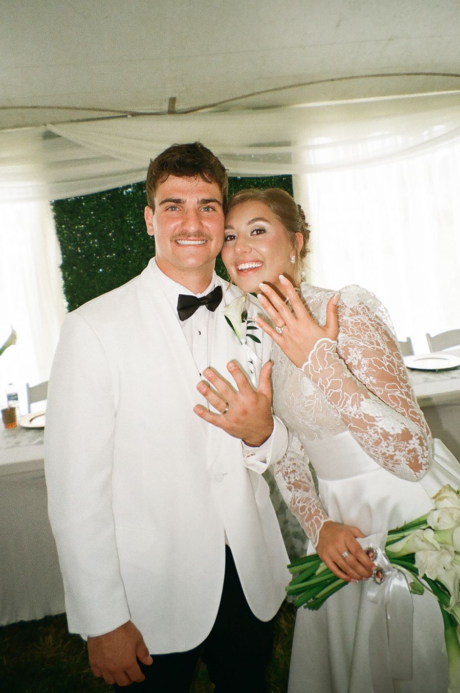 Smiling newlyweds in white wedding attire showing off their rings at an elegant tented reception venue.