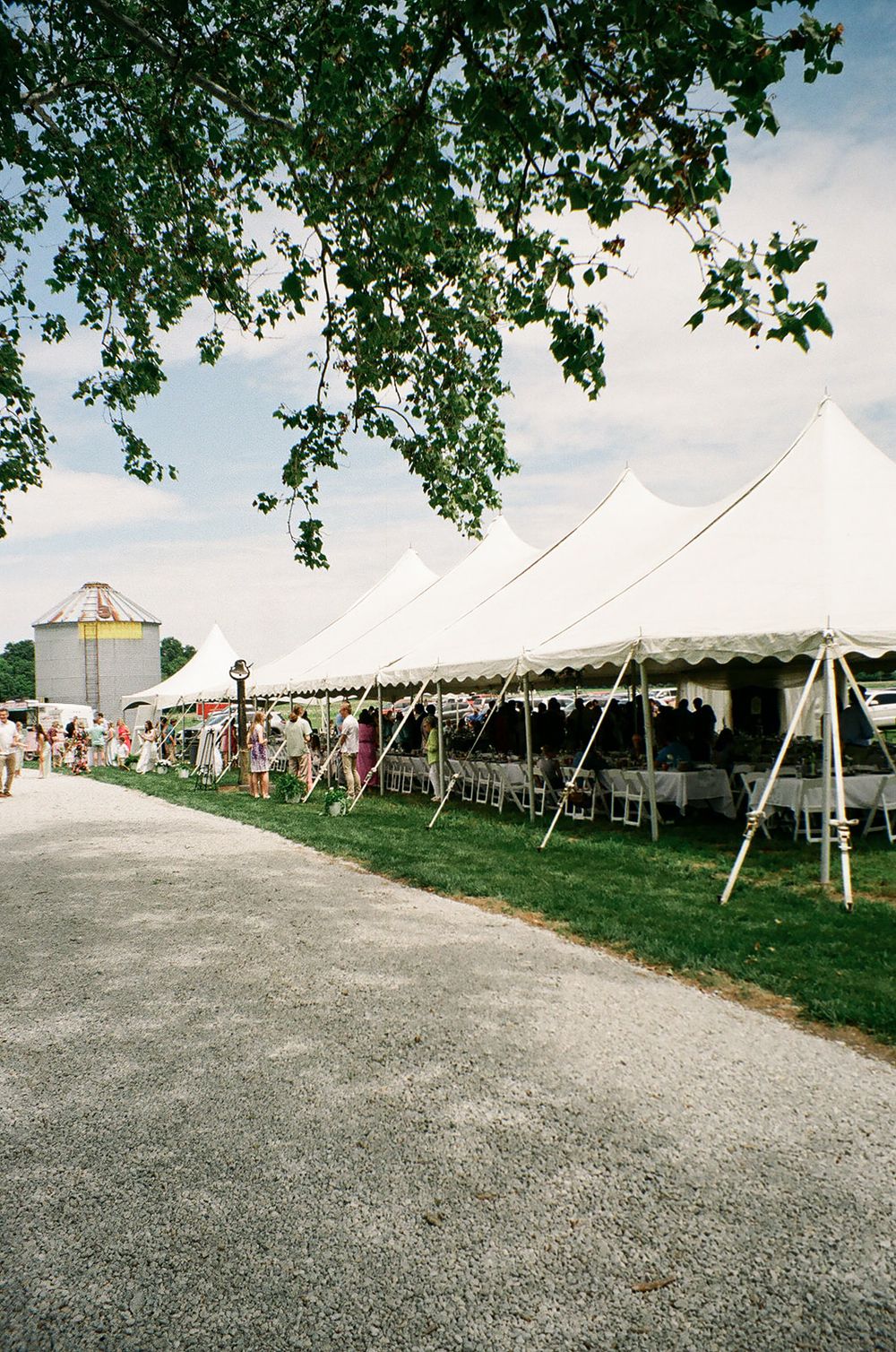 Large white event tent set up on green grass at an outdoor venue with trees and grain silo in background.