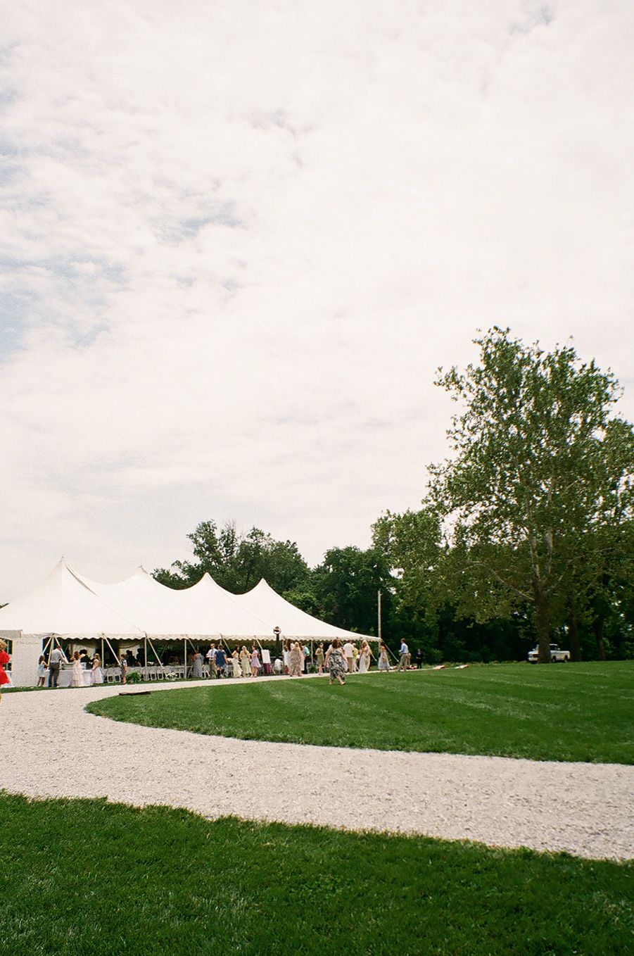 White wedding tent on lush green lawn with gravel path, trees, and guests gathered under cloudy sky.