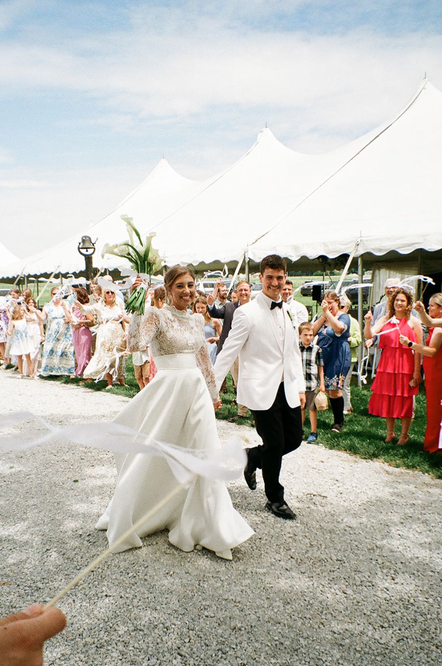 Bride in lace gown and groom in white tuxedo jacket walk joyfully through cheering guests at outdoor wedding reception.