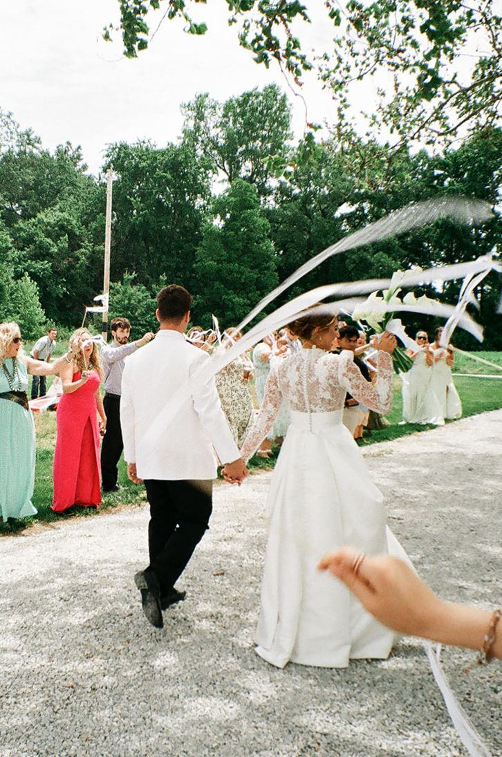 Bride and groom walk hand-in-hand through cheering guests waving white ribbons at an outdoor wedding celebration.