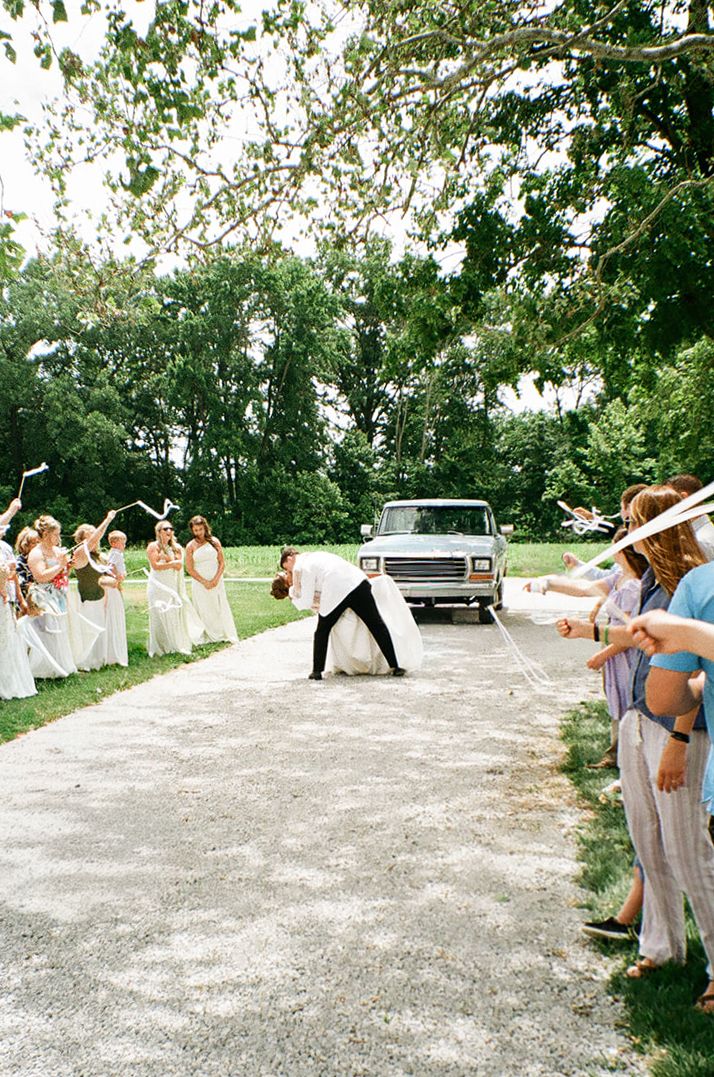 Bride and groom share a romantic dip kiss during outdoor wedding sendoff with guests lining gravel path waving streamers.