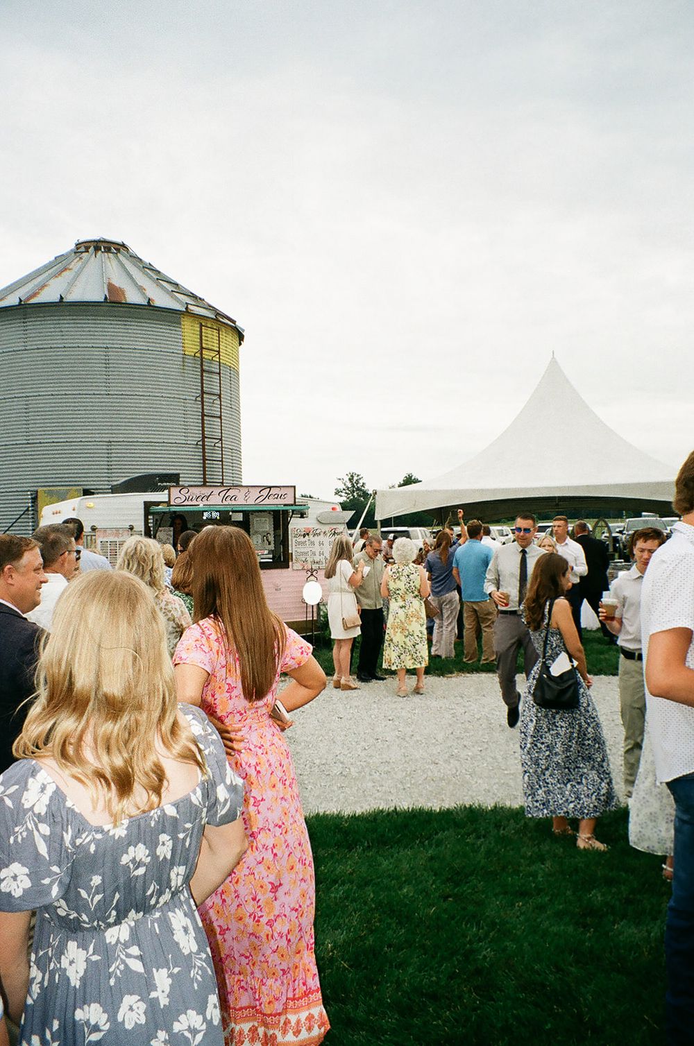 Guests mingle outdoors at a rustic farm wedding venue with a grain silo and white tent.