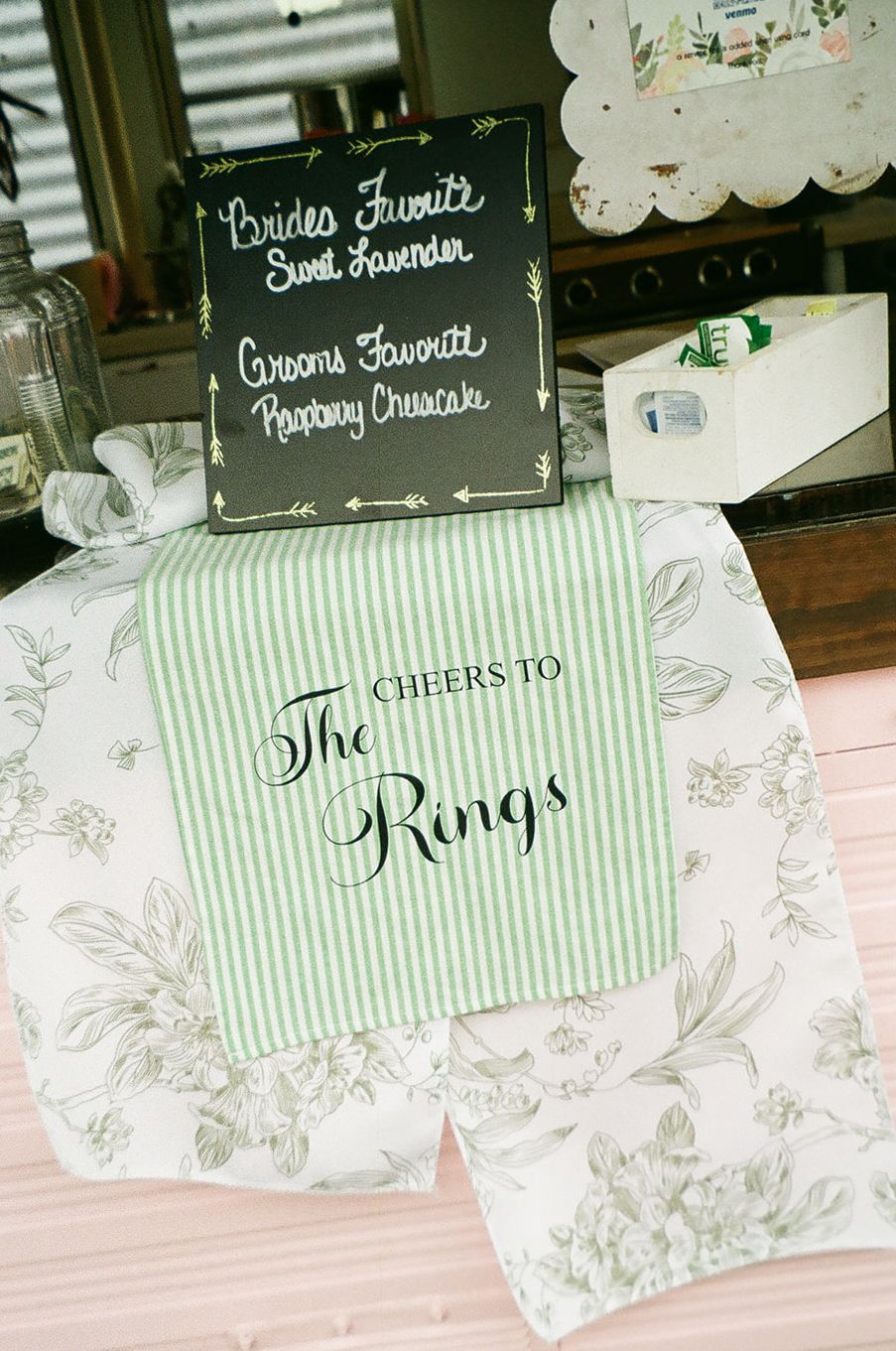 Green striped table runner with Cheers to The Rings text at wedding reception with chalkboard flavor sign.