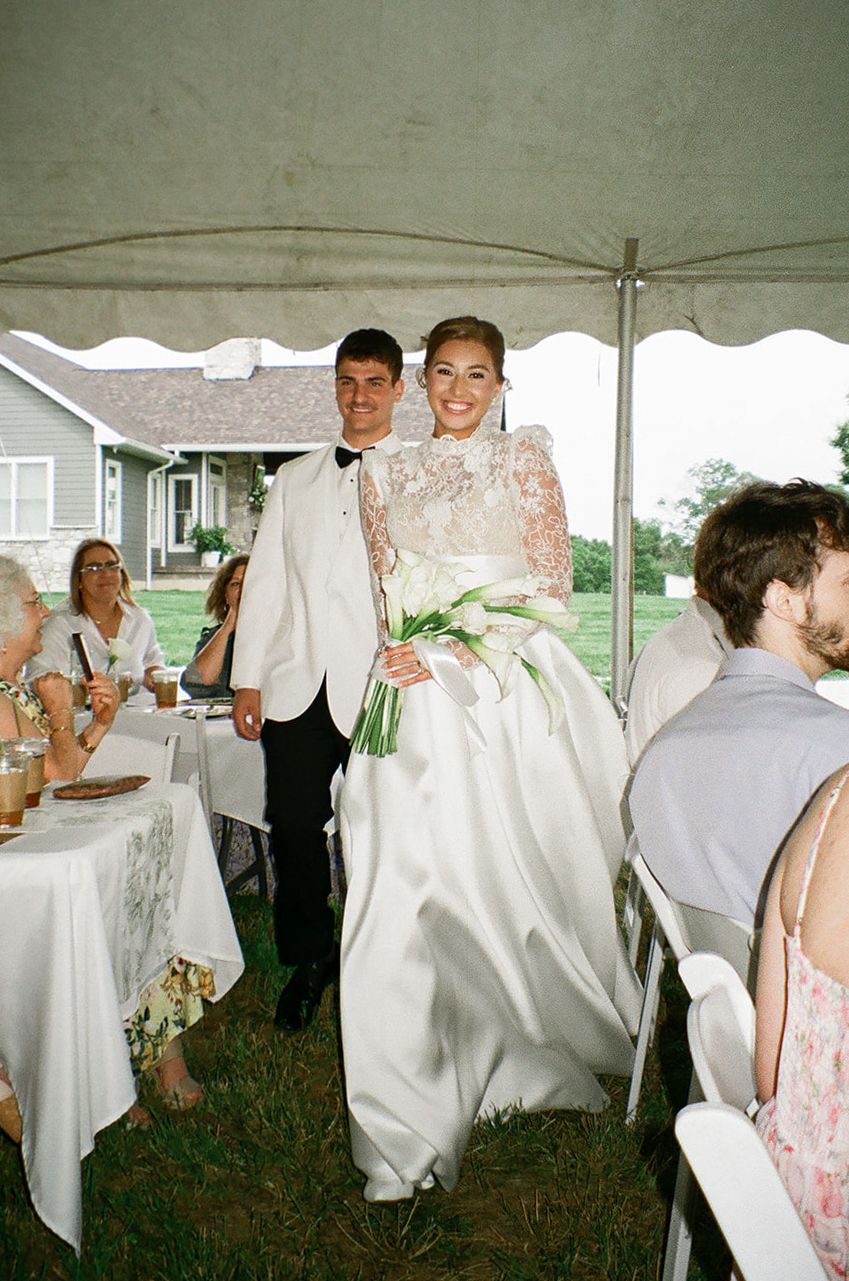 Bride in lace gown holding white bouquet walks with groom in white tuxedo at outdoor tent wedding.