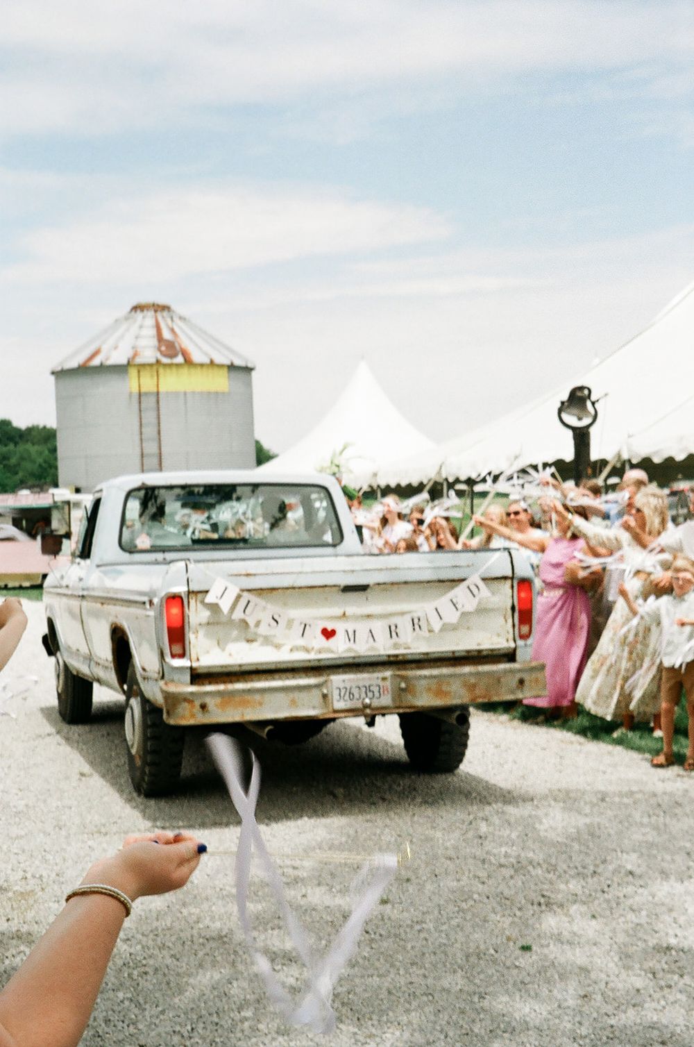 Just married couple drives away in rustic white pickup truck as wedding guests celebrate with streamers outdoors.