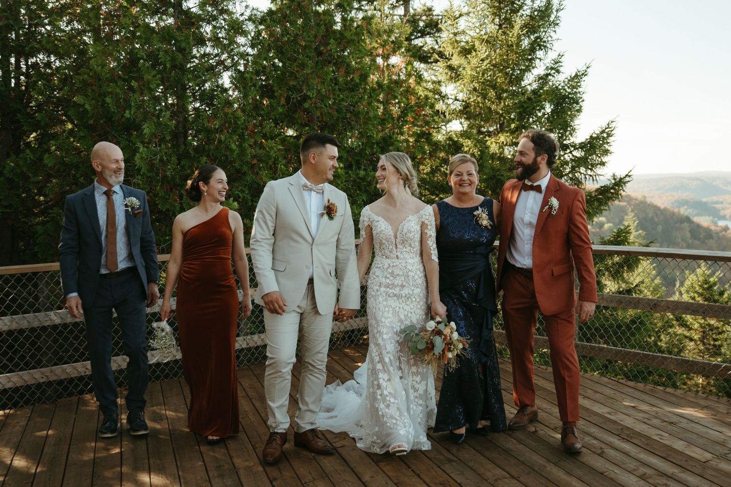 Wedding party posing on wooden deck with forest backdrop, bride in lace gown, groom in white suit, guests in formal attire.