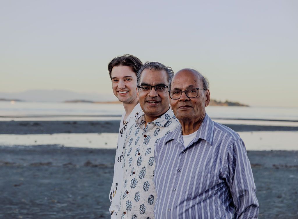 Three men of different generations pose together on a beach at sunset with calm water behind them.